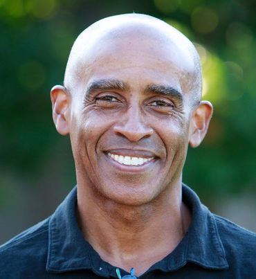 Smiling man with shaved head, wearing a black collared shirt, against a blurred green background.
