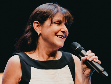 Woman with dark hair, smiling, holding a microphone. She wears a black and white dress on a stage.