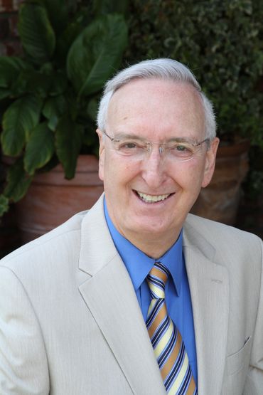 Man in light suit, blue shirt, and tie, smiling, with glasses, standing outside.