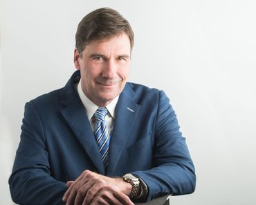 Man in a blue suit, smiling slightly, leaning on a chair against a white background.