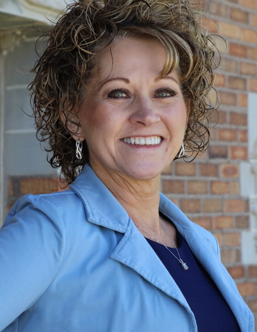 Woman smiling, wearing a blue blazer and top, with curly hair; brick background.