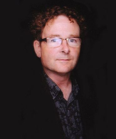Man with glasses and curly brown hair, wearing a patterned shirt and a black jacket, looking forward.