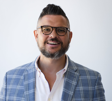 Man with glasses, beard, and pompadour haircut smiles, wearing a blue plaid blazer and pink shirt.
