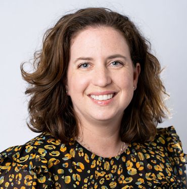 Woman smiling, wearing a black top with yellow and orange floral print, short dark hair, white background.