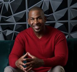 Man in red sweater, hands clasped, smiling; seated on a green couch against a geometric wall.
