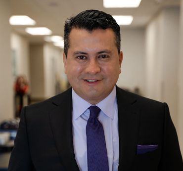 Man in suit smiles, standing in a brightly lit hallway. He wears a purple tie and has a pocket square.