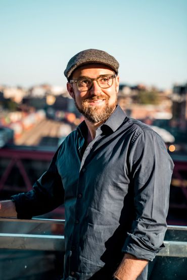 Man with glasses and a cap, smiling, leaning on a railing. City buildings in the background.