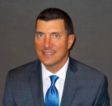 Man in a gray suit and blue tie smiles, against a gray background.