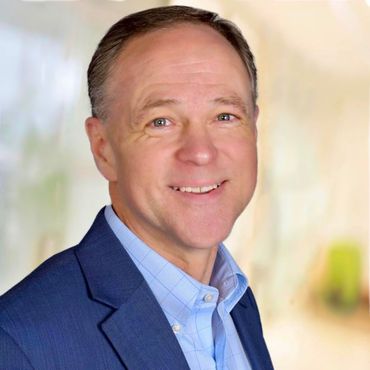 Man in blue blazer and shirt smiling, blurred background.