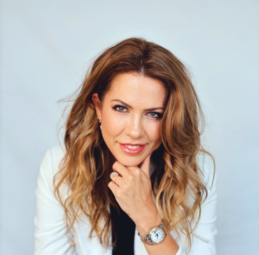 Woman with wavy brown hair, wearing a white blazer, resting chin on hand, smiling.