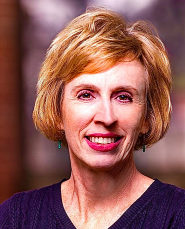 Woman with short blond hair smiles at the viewer, wearing a dark blue shirt; background blurred.