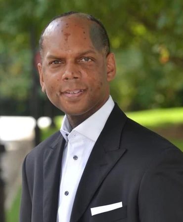 Man in black suit and white shirt smiles outdoors.