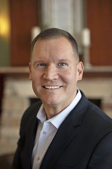 Man in a suit smiles at the camera, blurred background of a fireplace and candles.