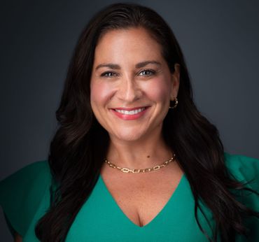 Woman with dark hair, smiling, wearing a green top and gold necklace against a dark gray background.