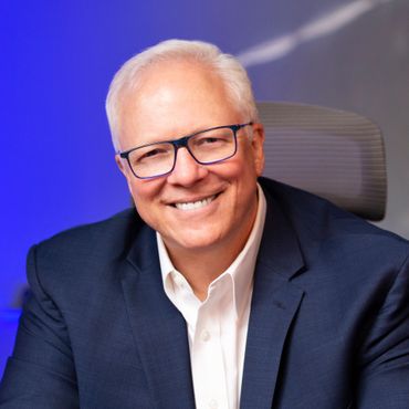 Smiling man with glasses wearing a blue blazer and white shirt, set against a blue background.