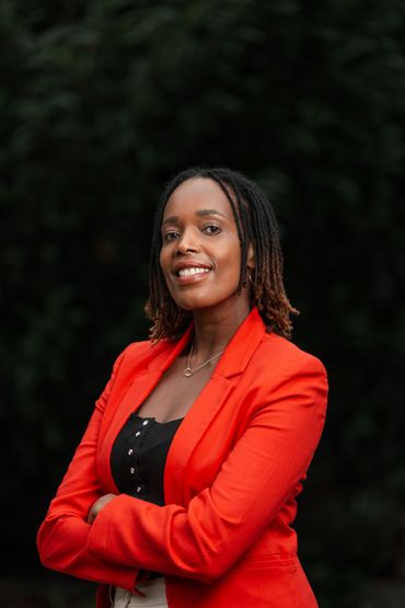 Woman in red blazer with arms crossed, smiling, set against a dark background.
