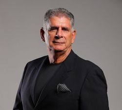 Man in a black blazer and shirt, looking at the camera. He's in front of a gray backdrop.