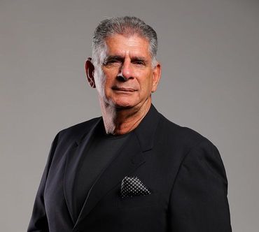 Man in a black blazer and shirt, looking at the camera. He's in front of a gray backdrop.