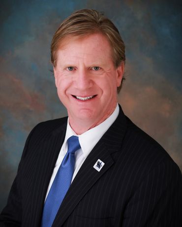 Man in suit smiling, with blue tie and dark blazer, against a blurred background.