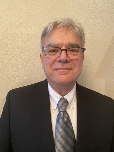 Man in glasses, suit, and tie, standing against a neutral-colored wall.