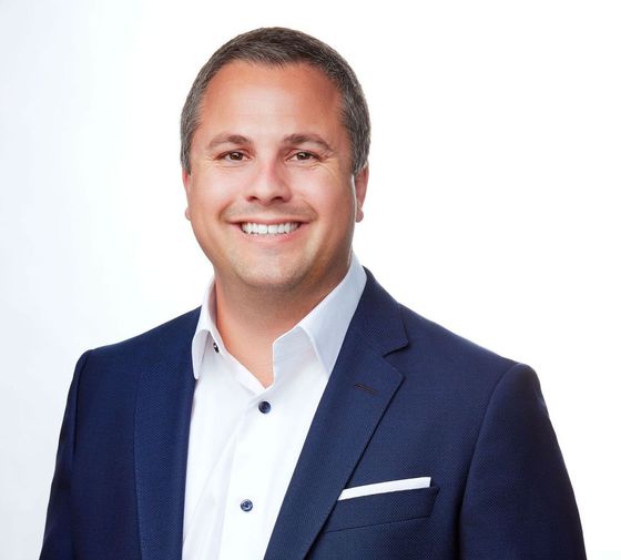 Man in a navy blazer and white shirt smiling at the camera against a white background.