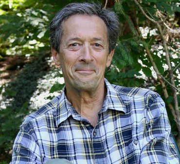 Man in plaid shirt smiles outdoors, surrounded by greenery.
