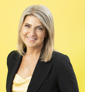 Woman with blonde hair, smiling, wearing a black blazer over a yellow top, against a yellow background.