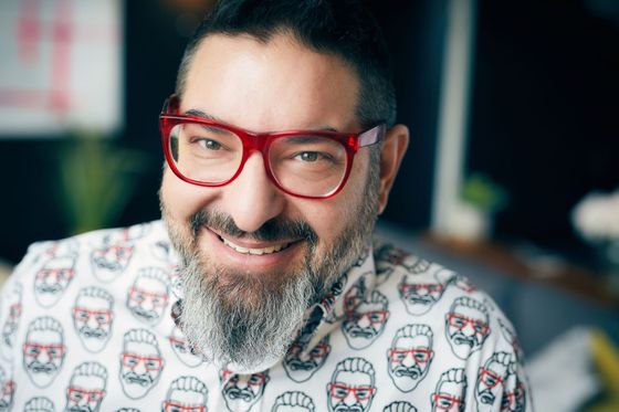 Man with a gray beard and red glasses smiles, wearing a white shirt with red face print.