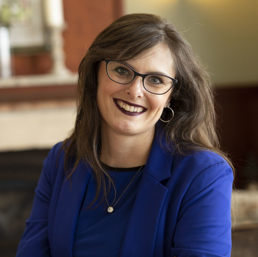 Woman with glasses smiles, wearing blue blazer and necklace, indoors.