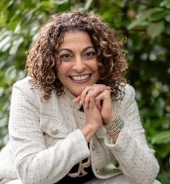 Woman with curly hair smiles, clasping hands, wearing a white jacket with a leafy green backdrop.