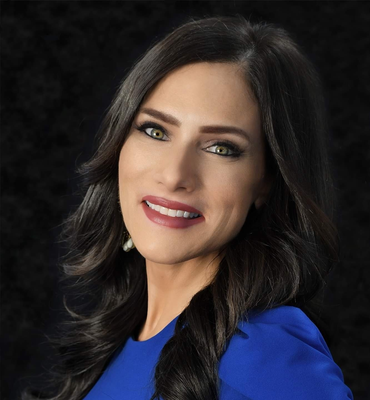 Woman with dark hair, green eyes, and a blue dress smiles against a black background.