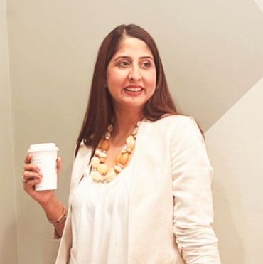 Woman smiling, holding a coffee cup, wearing a white outfit and necklace, standing indoors.