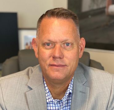 Man wearing a gray blazer and checkered shirt, seated, looking at the camera.