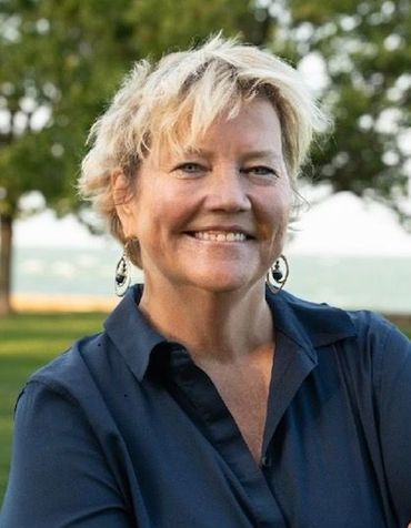Woman with short blonde hair smiles outdoors near water, wearing a dark blue collared shirt and earrings.