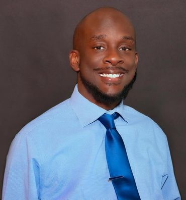 Man in blue shirt and tie smiles at the camera against a gray background.