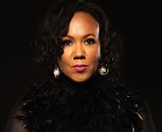 Woman with dark curly hair and a feathered top, wearing pearl earrings, looking forward.