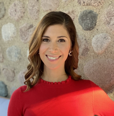 Woman with brown hair smiles, wearing a red sweater, in front of a stone wall.