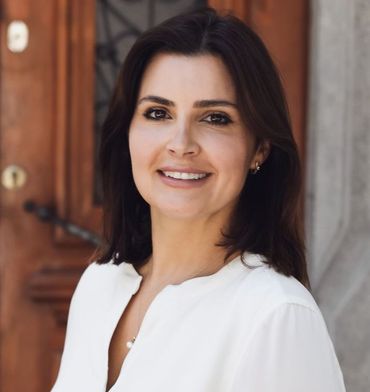 Woman with dark hair smiles, wearing a white blouse, standing in front of a wooden door.