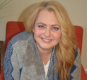 Woman with blonde hair wearing a patterned blue and gray sweater, smiling, seated in an orange chair.