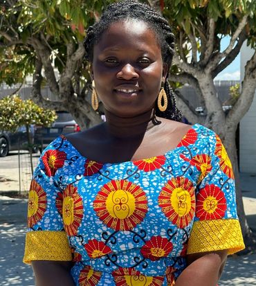 Woman in a blue patterned dress smiles, gold earrings, under a tree on a sunny day.