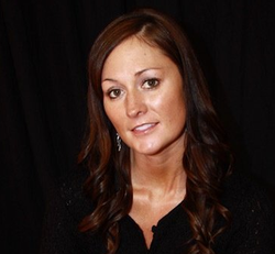 Woman with brown hair, wearing a black top, smiles slightly against a black backdrop.