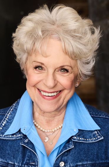 Woman with short, curly, white hair smiles, wearing a blue denim jacket over a light blue shirt.