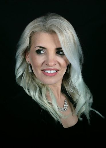 Woman with long blonde hair, smiling, wearing a necklace and earrings, against a black background.