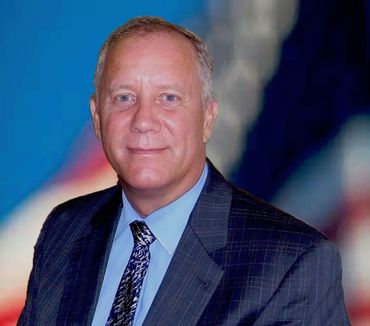 Man in a dark suit and tie, smiling, against a blurred blue background.