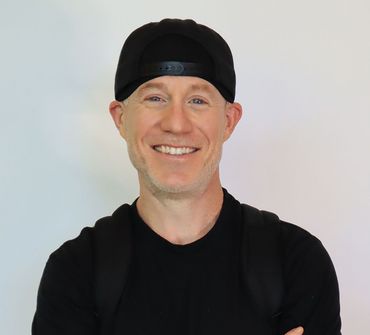 Smiling person wearing a black cap and shirt, with a backpack against a white backdrop.