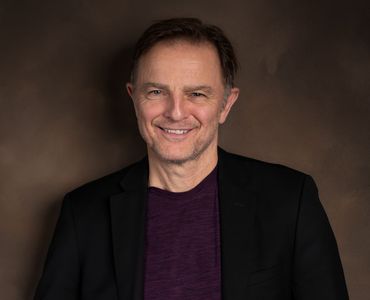 Man smiling, leaning against a brown wall. He wears a black blazer and purple shirt.