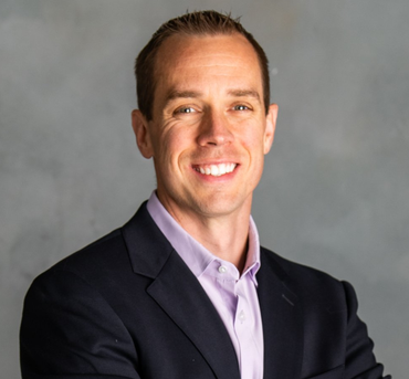 Smiling man in a dark suit jacket, light purple shirt, against a gray background.
