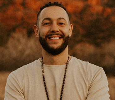 Smiling person with a beard, wearing a beige shirt and beaded necklace, set against a blurred outdoor background.