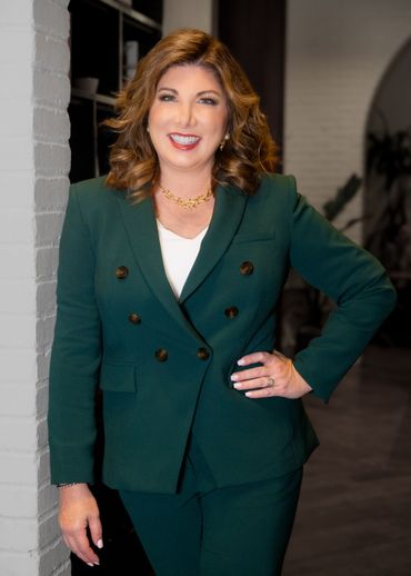 Woman in a green suit, smiling, standing in an office with a brick wall and shelving.
