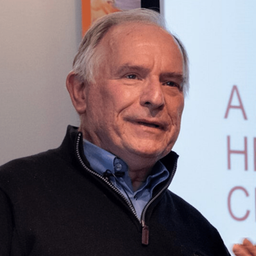 Man in navy sweater and blue shirt speaks in front of a screen.